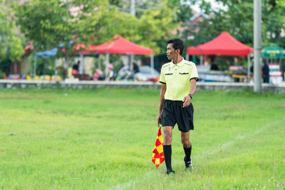 Assistant referee in bright uniform carrying flag on a grassy football field during a match day.